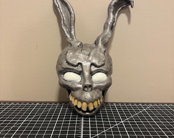 May include: A silver-colored mask in the shape of a rabbit's head with a human-like skull. The mask has large, pointed ears, white eye sockets, and yellow teeth. The mask is likely a prop or decorative item.
