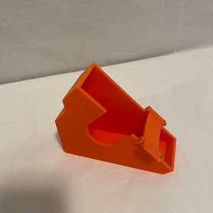 Puede incluir: Soporte para teléfono de plástico naranja con una ranura para un teléfono y un borde elevado para evitar que el teléfono se deslice. El soporte está diseñado para sostener un teléfono en posición vertical.