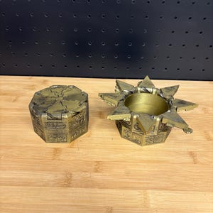 Può includere: Scatola puzzle dorata in due pezzi con disegni geometrici e simboli simili a geroglifici. La scatola è aperta, rivelando l'interno. La scatola è realizzata in materiale metallico e poggia su una superficie di legno.