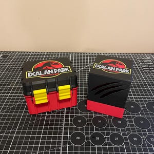 May include: Two black boxes with red and yellow accents. The boxes have a Jurassic Park logo with the text "DXALAN PARK" on the top. One box has a claw mark design on the front.