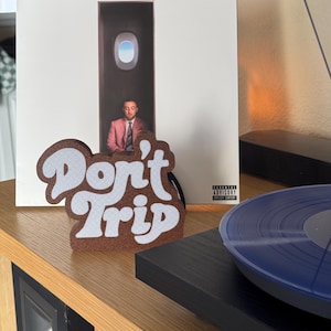 Pode incluir: Uma decora&ccedil;&atilde;o de feltro castanho e branco com as palavras "Don't Trip" em uma fonte retr&ocirc;. A decora&ccedil;&atilde;o est&aacute; em frente a um &aacute;lbum de vinil com um homem de terno rosa. Um disco de vinil azul est&aacute; em uma vitrola.