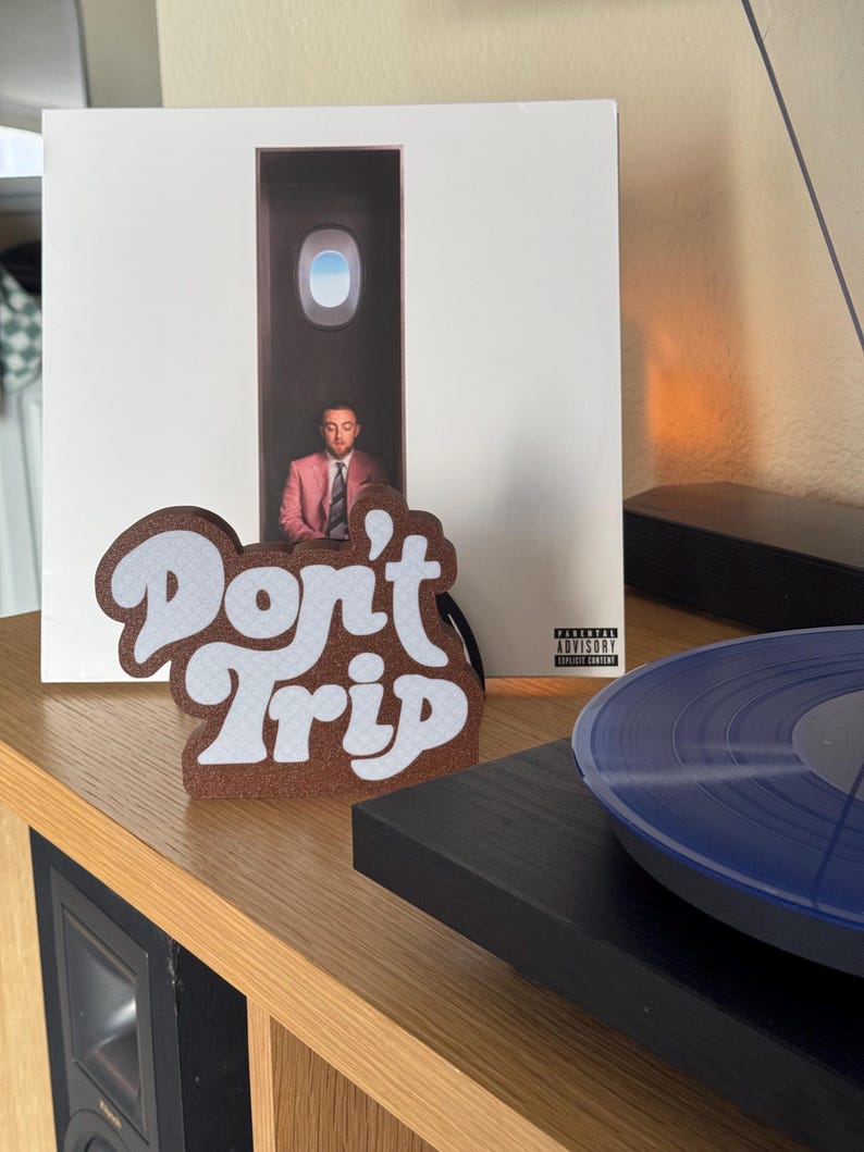 Pode incluir: Uma decora&ccedil;&atilde;o de feltro castanho e branco com as palavras "Don't Trip" em uma fonte retr&ocirc;. A decora&ccedil;&atilde;o est&aacute; em frente a um &aacute;lbum de vinil com um homem de terno rosa. Um disco de vinil azul est&aacute; em uma vitrola.