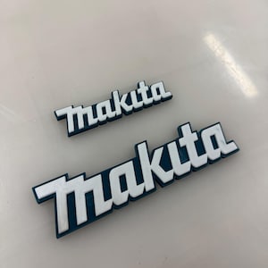 May include: Two white plastic Makita logos on a white surface. The larger logo is teal blue and the smaller logo is a lighter shade of teal blue.