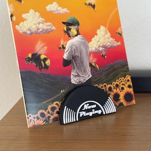 Puede incluir: Un soporte para discos de vinilo con un diseño en blanco y negro. El soporte tiene forma de disco y tiene las palabras "Now Playing" escritas en él. El soporte está sosteniendo un disco de vinilo con una imagen colorida de un hombre con abejas y girasoles.
