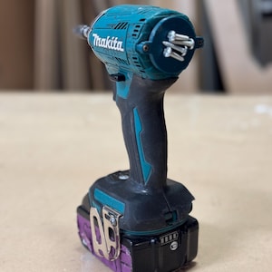 Puede incluir: Un taladro inalámbrico de color azul verdoso y negro con una batería. El taladro tiene un porta puntas magnético con varios tornillos unidos a él. La marca "Makita" es visible en el taladro.