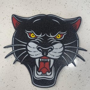 May include: Black panther head with a white snout and red mouth, with yellow eyes. The panther is depicted in a traditional tattoo style.