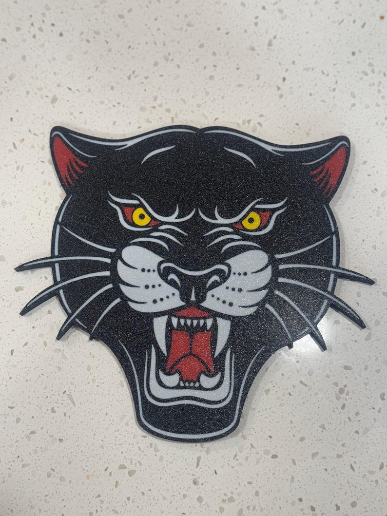 May include: Black panther head with a white snout and red mouth, with yellow eyes. The panther is depicted in a traditional tattoo style.