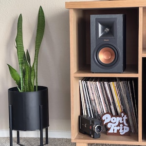 Pode incluir: Um alto-falante preto com um cone cor de cobre est&aacute; em uma prateleira de madeira ao lado de discos de vinil. Uma floreira preta com uma planta-cobra e uma c&acirc;mera vintage tamb&eacute;m s&atilde;o vis&iacute;veis. Uma placa que diz "Don't Trip" tamb&eacute;m est&aacute; na prateleira.