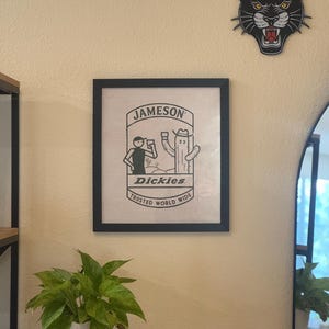 May include: A black framed print with a white background featuring a cartoon illustration of a person and a cactus drinking from glasses. The text "Jameson" and "Dickies" are printed in black. The text "Trusted World Wide" is printed in black below the illustration.
