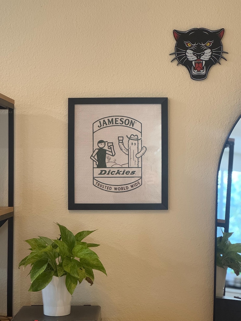 May include: A black framed print with a white background featuring a cartoon illustration of a person and a cactus drinking from glasses. The text "Jameson" and "Dickies" are printed in black. The text "Trusted World Wide" is printed in black below the illustration.