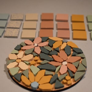 May include: A round mosaic tile coaster with a floral design. The coaster features pink, yellow, green, and blue tiles arranged in a flower pattern. The coaster is surrounded by small square tiles in various shades of blue, beige, pink, yellow, and green.