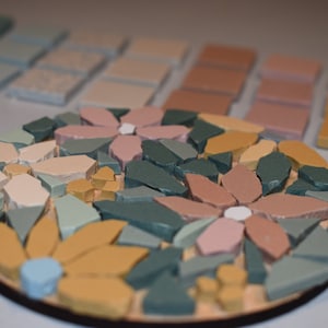 May include: A round mosaic tile coaster with a floral design. The coaster features pink, yellow, and green flowers with white centers. The coaster is made of small, irregular pieces of tile.