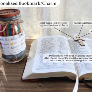 May include: A personalized bookmark and charm set. The image shows a wooden cross bookmark with a ribbon, resting on an open book. A jar filled with colorful sticks and a personalized tag are also visible. The bookmark is engraved with a name and can be customized.