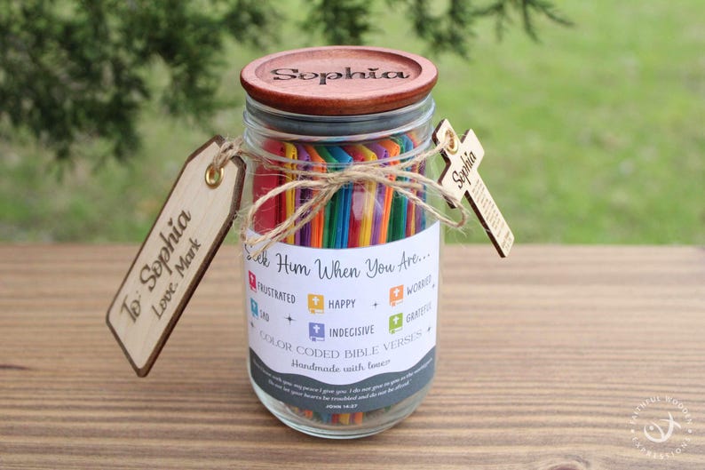 May include: A glass jar with a wooden lid engraved with the name "Sophia". The jar contains colorful sticks and is adorned with wooden tags and a cross. The jar has a label with the text "Seek Him When You Are..." and "Color Coded Bible Verses".