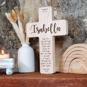 May include: A wooden cross with the name "Isabella" engraved on it. The cross also has a Bible verse from Romans 15:13 engraved on it. The cross is sitting on a wooden surface with a rainbow toy and a vase with dried flowers in the background.