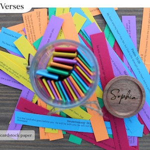 May include: A collection of colorful paper strips with Bible verses, scattered around a glass jar filled with colorful sticks. A wooden coaster with the name "Sophia" is also present. The text "Over 100 Verses" and "Heavier 65lb cardstock paper" are visible.