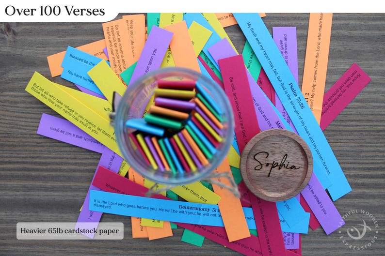 May include: A collection of colorful paper strips with Bible verses, scattered around a glass jar filled with colorful sticks. A wooden coaster with the name "Sophia" is also present. The text "Over 100 Verses" and "Heavier 65lb cardstock paper" are visible.