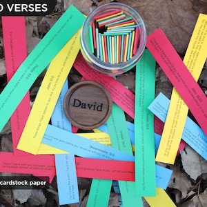 May include: A display of colourful paper strips with Bible verses, arranged amongst dried leaves. A small wooden disc with the name "David" is visible, alongside a glass jar filled with colourful sticks. Text on the image reads "OVER 100 VERSES" and "Heavier 65lb cardstock paper."
