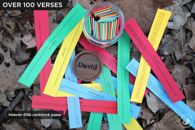 May include: A display of colourful paper strips with Bible verses, arranged amongst dried leaves. A small wooden disc with the name "David" is visible, alongside a glass jar filled with colourful sticks. Text on the image reads "OVER 100 VERSES" and "Heavier 65lb cardstock paper."