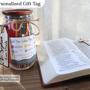 May include: A glass jar filled with colorful pencils, a wooden lid, and a personalized gift tag. The tag reads "To: Sophia Love, Mark." The jar is next to an open book with a wooden cross.