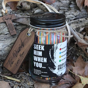 May include: A glass jar with a black lid and a label that reads "Seek Him When You..." contains colorful sticks. A wooden cross and a tag with the name "David" are attached. The jar is tied with twine and sits on a bed of leaves.