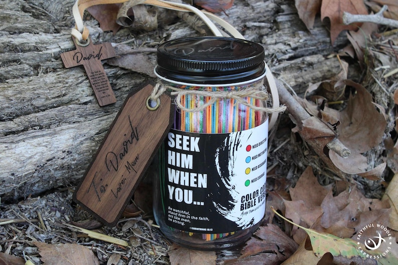 May include: A glass jar with a black lid and a label reading "Seek Him When You..." contains colourful sticks. A wooden cross and a tag with the name "David" are attached. The jar is tied with twine and rests on a bed of leaves.