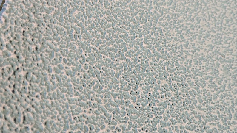 May include: Close-up of a textured surface with a pattern of small, irregular bumps. The surface has a light blue-green color with a slightly off-white background, creating a mottled appearance. The texture is rough and uneven.
