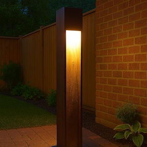 May include: A tall, rectangular outdoor light fixture with a warm, glowing interior. The dark brown metal structure stands on a brick patio near a wooden fence and a brick wall. The light illuminates the surrounding area.