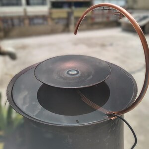 May include: A copper-colored water fountain with a curved spout and a circular basin. The fountain has a dark, metallic finish and a modern design. The water feature is outdoors, with a blurred background.