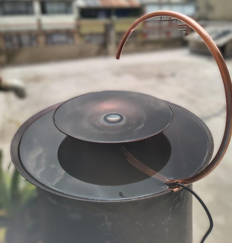 May include: A copper-colored water fountain with a curved spout and a circular basin. The fountain has a dark, metallic finish and a modern design. The water feature is outdoors, with a blurred background.