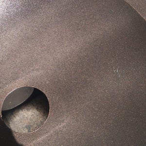May include: Close-up of a dark brown, textured metal surface with a circular hole. The surface has a slightly rough, speckled appearance, and the hole is smooth and dark.