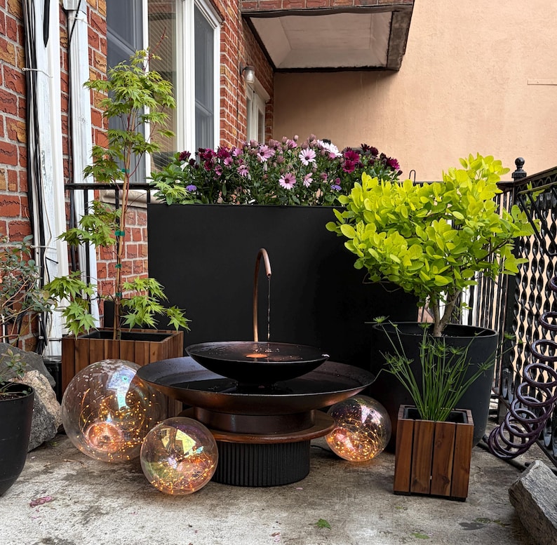 May include: Outdoor water fountain with a copper spout and a black basin. The fountain is surrounded by plants in wooden and black pots, with decorative glass orbs lit with warm lights. A black planter box holds colorful flowers.