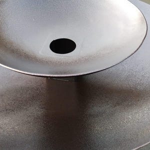 May include: A two-tiered, metallic brown decorative sculpture with a textured surface. The top tier is a bowl shape with a central hole, set atop a larger, flat, circular base. The sculpture has a modern, minimalist design.