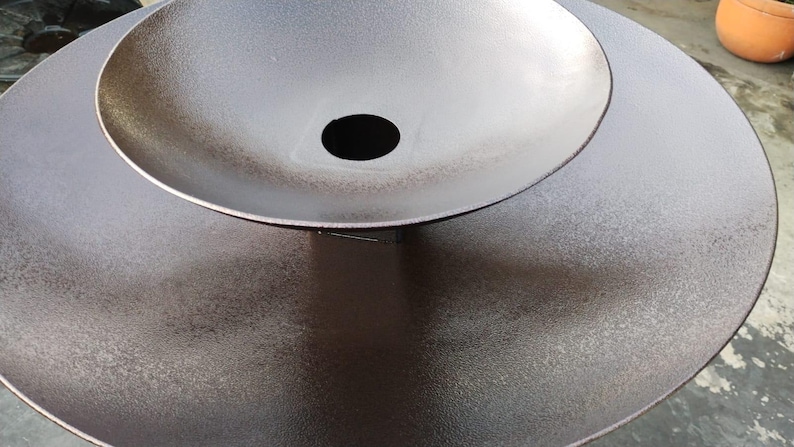 May include: A two-tiered, metallic brown decorative sculpture with a textured surface. The top tier is a bowl shape with a central hole, set atop a larger, flat, circular base. The sculpture has a modern, minimalist design.
