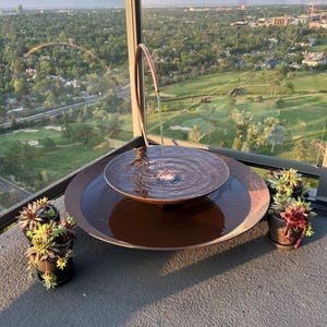 May include: A tiered copper-colored metal water fountain with a curved spout pouring water into a central basin, which overflows into a larger circular base. Two small dark pots containing green and reddish succulent plants sit on either side.