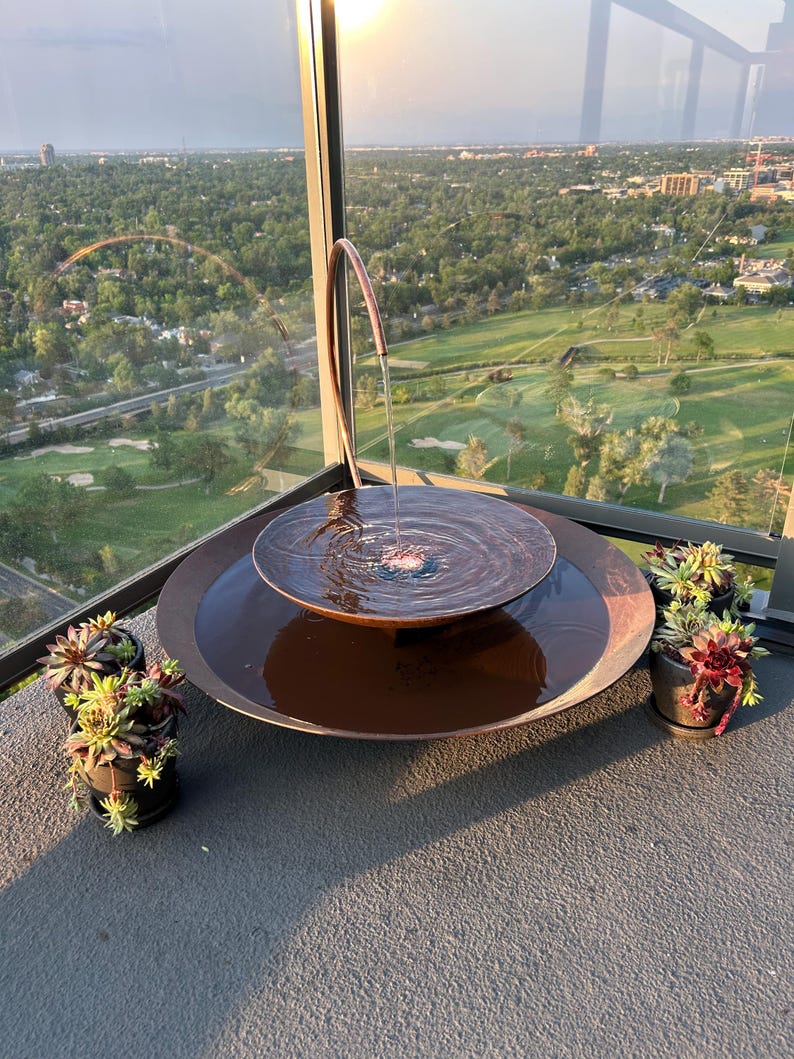 May include: A tiered copper-colored metal water fountain with a curved spout pouring water into a central basin, which overflows into a larger circular base. Two small dark pots containing green and reddish succulent plants sit on either side.