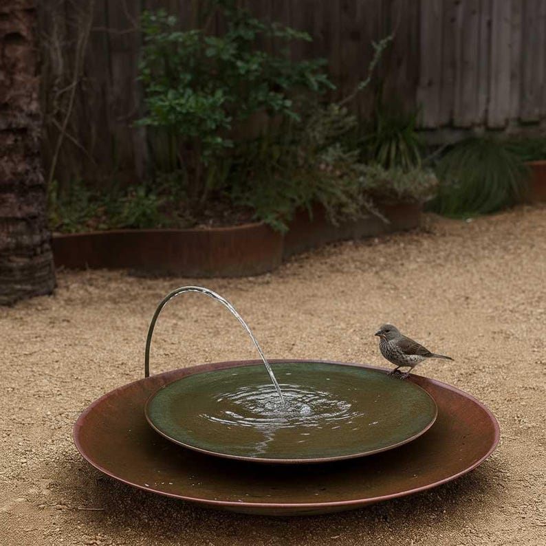 May include: A tiered outdoor water fountain with a dark green basin and a bronze-colored base. A small bird perches on the edge of the basin. Water arcs from a curved spout into the basin, creating ripples.