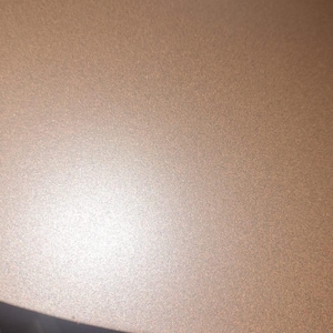 May include: Close-up of a section of a metallic, textured surface. The color is a warm, muted bronze or copper tone, with a subtle sheen. The surface appears to be slightly curved, with a darker, black area at the bottom.