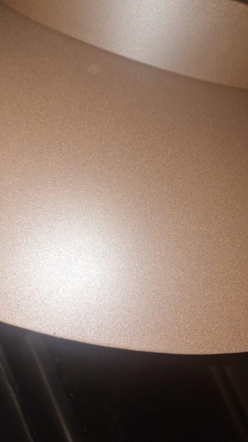May include: Close-up of a section of a metallic, textured surface. The color is a warm, muted bronze or copper tone, with a subtle sheen. The surface appears to be slightly curved, with a darker, black area at the bottom.