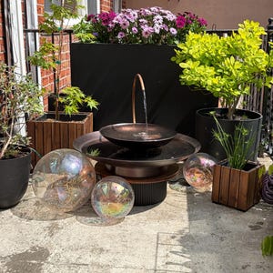 May include: Outdoor water fountain with a copper-colored bowl and a dark base. The fountain is surrounded by potted plants, including a small tree and flowering plants. Iridescent glass spheres are placed around the fountain.