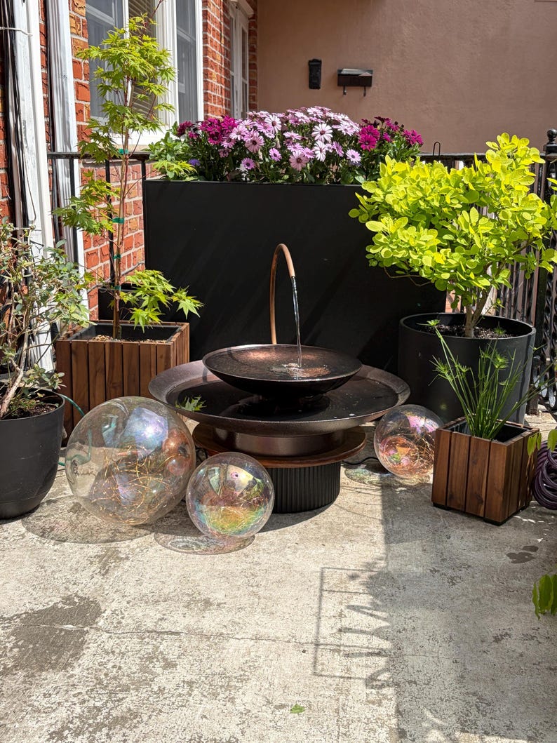 May include: Outdoor water fountain with a copper-colored bowl and a dark base. The fountain is surrounded by potted plants, including a small tree and flowering plants. Iridescent glass spheres are placed around the fountain.