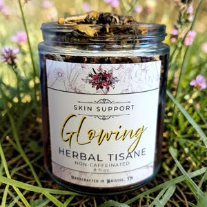 May include: A glass jar filled with herbal tisane, labeled "Glowing Herbal Tisane" for skin support. The label includes the text "Handcrafted in Bristol, TN" and "8 fl.oz.". The jar is set against a backdrop of green grass and purple flowers.