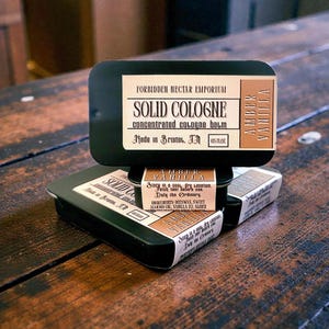 Puede incluir: Tres latas rectangulares negras de colonia sólida apiladas sobre una superficie de madera. La lata superior tiene una etiqueta que dice "Amber Vanilla" y "Solid Cologne". También es visible el texto "Forbidden Nectar Emporium" y "Made in Bristol, TN".