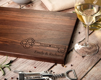 Puede incluir: Una tabla de cortar de madera marrón con un diseño de llave grabado en ella. La llave tiene el texto "New Home New Adventures 2024" y los nombres "Emily & Richard" están grabados debajo de la llave.