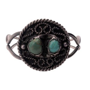 May include: A silver cuff bracelet featuring a circular centerpiece with a twisted rope border and intricate wire scrollwork. Two oval-shaped turquoise stones are set side-by-side in the center of the dark, oxidized silver metal design.