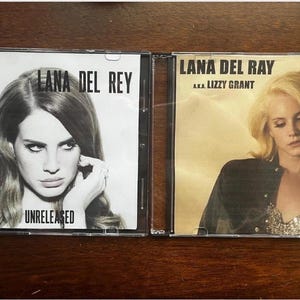 May include: Two CD cases featuring Lana Del Rey album covers. One is black and white with the text "Lana Del Rey" and "Unreleased." The other has a golden hue with the text "Lana Del Rey a.k.a. Lizzy Grant."