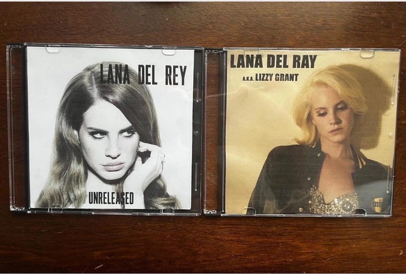 May include: Two CD cases featuring Lana Del Rey album covers. One is black and white with the text "Lana Del Rey" and "Unreleased." The other has a golden hue with the text "Lana Del Rey a.k.a. Lizzy Grant."