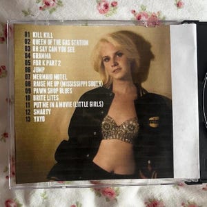 May include: A CD case featuring a woman with blonde hair and a black jacket. The tracklist includes titles like "Kill Kill," "Queen of the Gas Station," and "Mermaid Motel." The case is in a clear plastic sleeve.