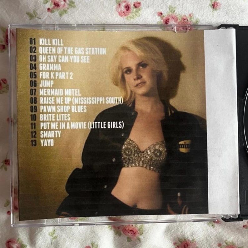 May include: A CD case featuring a woman with blonde hair and a black jacket. The tracklist includes titles like "Kill Kill," "Queen of the Gas Station," and "Mermaid Motel." The case is in a clear plastic sleeve.