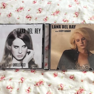 May include: Two CD cases featuring Lana Del Rey. One case has the text "Lana Del Rey" and "Unreleased" with a black and white photo. The other case says "Lana Del Rey a.k.a. Lizzy Grant" with a photo of the artist.